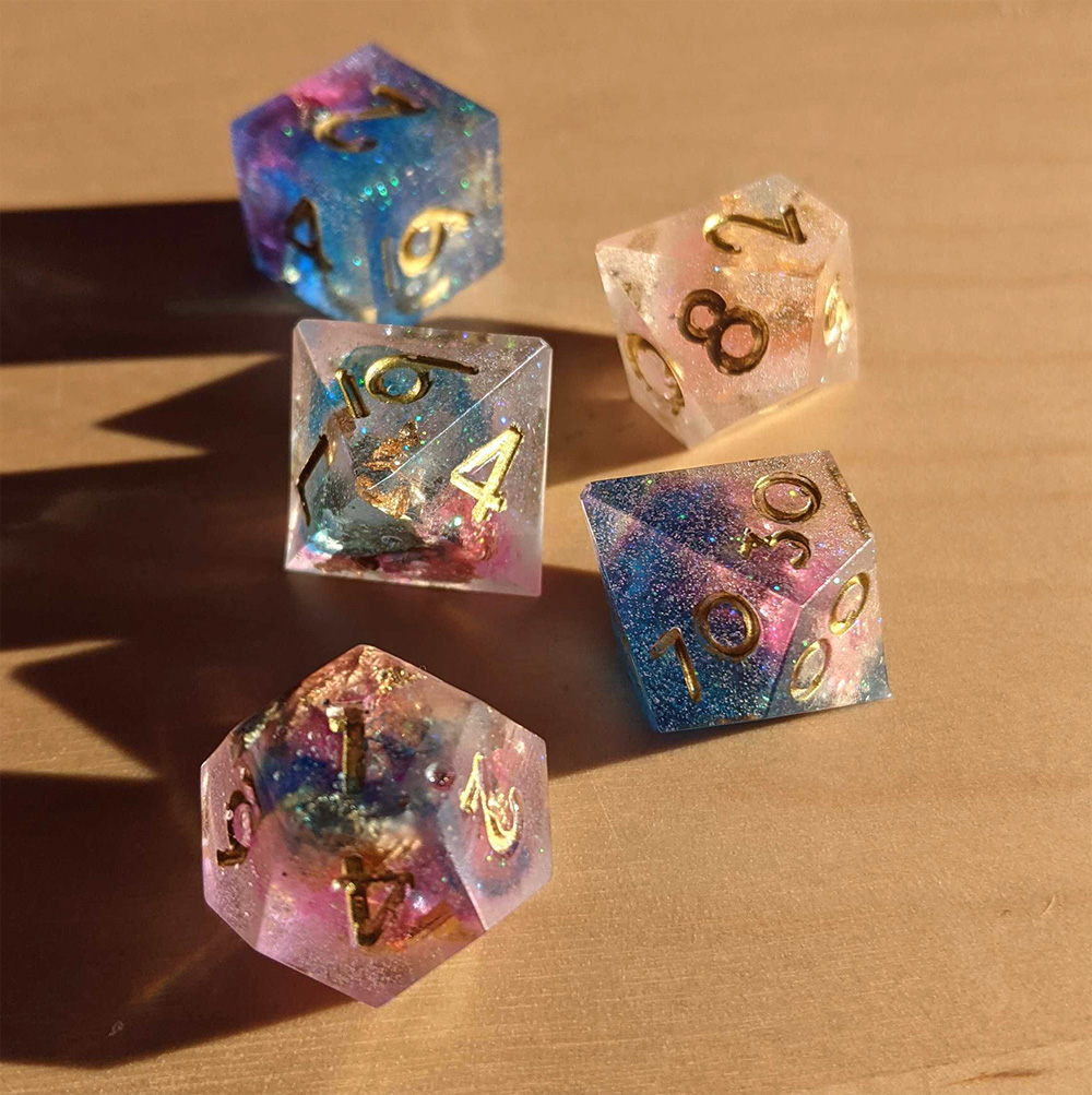 Galaxy-inspired dice, with blues, purples, pinks, and transparent body. The dice are inked with gold. With flecks of sparkles inside, it really looks like there's little galaxies in each!